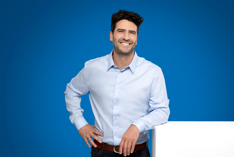 Um homem de camisa apoia-se com o braço numa caixa branca e olha sorridente para a câmara