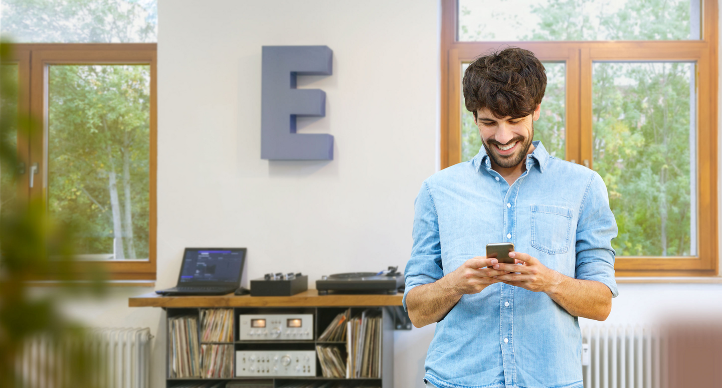 Um jovem de camisa azul está numa sala de estar, a sorrir e a olhar para o seu smartphone